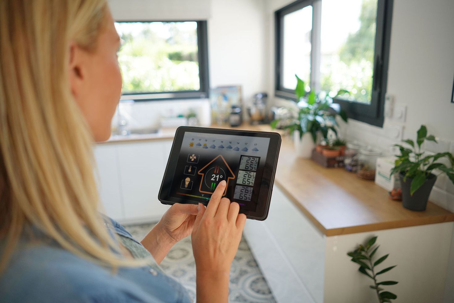 Eine Frau steht in einer Küche und hält ein Tablet, auf welchem über eine App das Wetter zu sehen ist, ebenso ist die Steuerung von Temperatur, Licht und Belüftung möglich