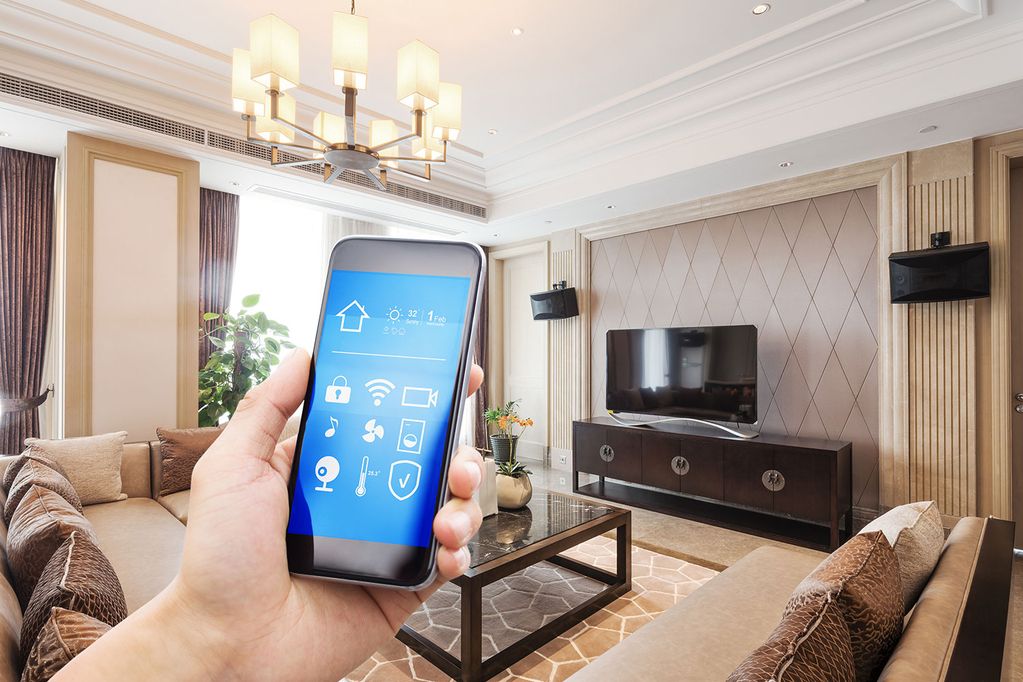 Eine Hand hält ein Handy mit geöffneter Smarthome App, im Hintergrund ist ein Wohnzimmer mit Sofa, Tisch, Kommode und Fernseher zu sehen